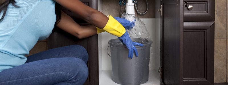 woman wearing rubber gloves with a bucket below a sink that's leaking water.